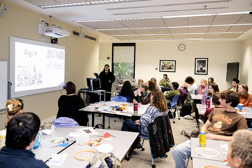 Students sit at desks with sewing supplies laid out on them, listening to the opening presentation of the workshop