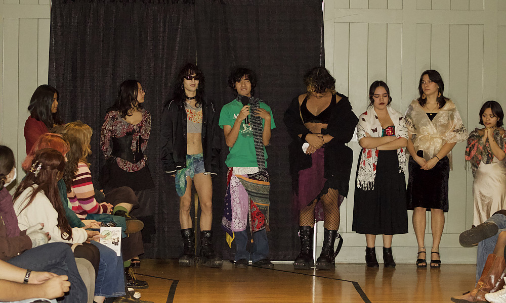 Fashion show participants stand in front of audience members, one holding a microphone and speaking
