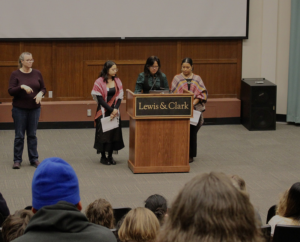 Three symposium co-chairs standing at the posium in Council Chamber, and an ASL interpreter nearby