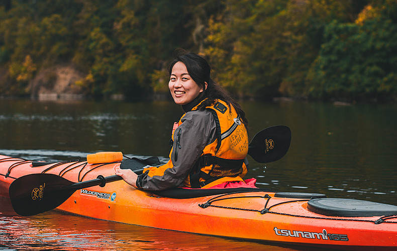A student smiles at the camera while in a kayak on water.