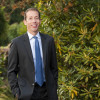 Bruce posing outside, wearing a dark suit and a blue tie.