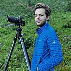 Nicholas standing outside in a blue jacket, next to a camera on a tripod.