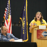 Student debating at a lecturn while a person sits at a table next to them.