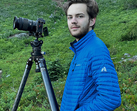 Nicholas standing outside in a blue jacket, next to a camera on a tripod.