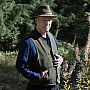 Kim Stafford posing outside, wearing a brimmed hat, green vest, and long sleeved blue shirt.