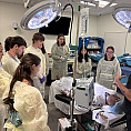 Group of students wearing surgical gear and standing by a dummy on a bed.