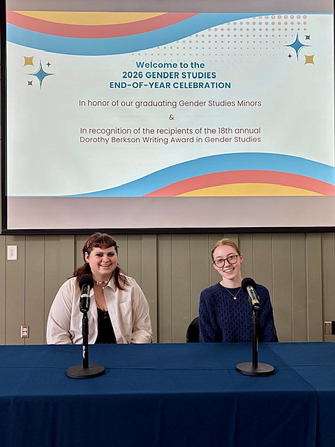 2 students sitting at table with microphones and large screen in background