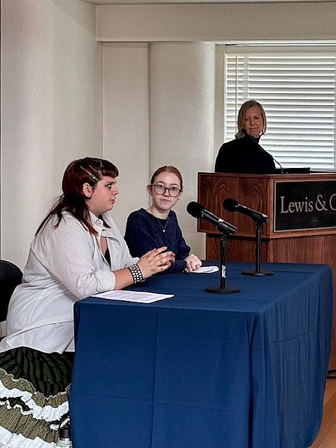 two students seated at a table with microphones and a person at a podium looking on