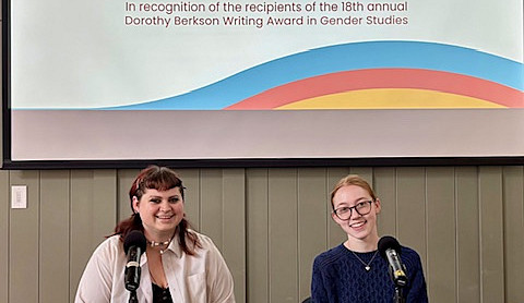 2 students sitting at table with microphones and large screen in background