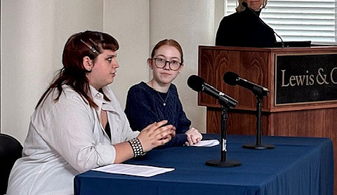 two students seated at a table with microphones and a person at a podium looking on